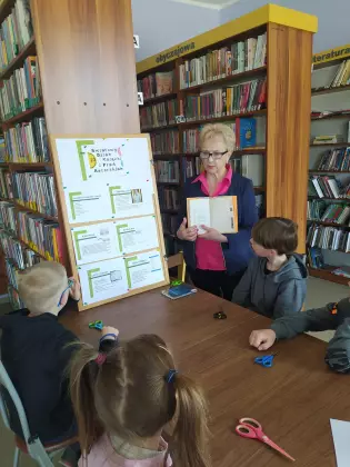 Uczniowie w bibliotece słuchają ciekawostek na temat książki opowiadanej przez Panią bibliotekarkę.