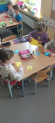 Przedszkolaki siedzą przy stolikach i wykonują pracę plastyczną.