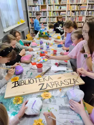 Dzieci na zajęciach malują kamienie i napisz przed bibliotekę na kawałku drewna.