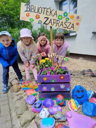 Dzieci stoją przed biblioteką na ziemi leżą pomalowane kamienie i drewniana doniczka z bratkami.