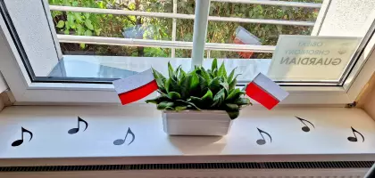 Na parapecie stoi doniczka z kwiatami i dwiema flagami Polski, obok są porozkładane czarne nuty z papieru.