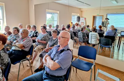 W sali na krzesłach siedzą seniorzy i słuchają występu górala grającego na akordeonie.