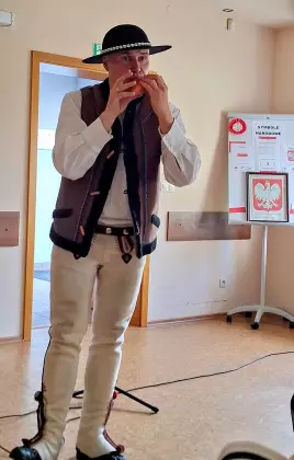 Góral Szczepan grający na okarynie, w tle tablica z godłem Polski i plakatami „Poznaj bliżej polskie symbole narodowe.