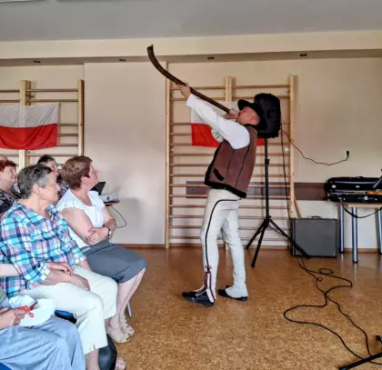 Góral Szczepan gra na trombicie, a seniorzy siedzący na krzesłach go słuchają.
