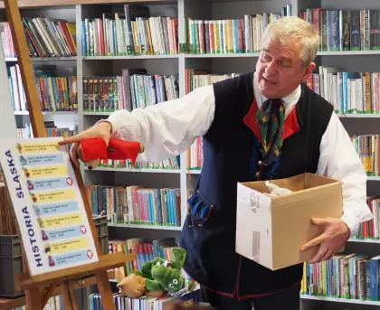 Pan Marek Szołtysek pokazuje planszę i trzyma karton, w tle regały z książkami.