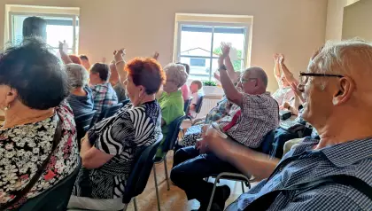 Seniorzy siedzą na krzesłach i z radością podnoszą ręce do góry wykrzykując góralskie Hej!.