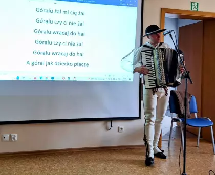 Góral Szczepan gra na akordeonie. W tle widać ekran z tekstem piosenki.