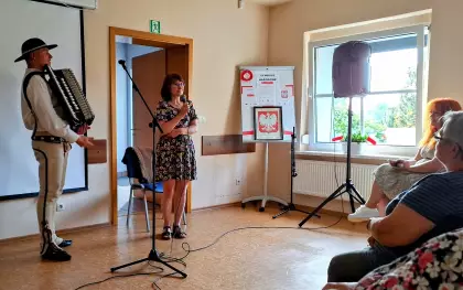 Przy mikrofonie stoi Góral Szczepan z akordeonem, a obok do mikrofonu mówi Pani Dyrektor Gminnej Biblioteki Publicznej w Pawonkowie. Na krzesłach siedzą ludzie i słuchają. W rogu sali stoi głośnik i tablica z godłem Polski i plakatami Poznaj bliżej polskie symbole narodowe.