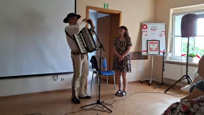W sali przed mikrofonem stoi mężczyzna w góralskim stroju i trzyma akordeon, obok stoi  Pani Dyrektor Gminnej Biblioteki Publicznej w Pawonkowie. W tle tablica z godłem Polski i plakatami „Poznaj bliżej polskie symbole narodowe.
