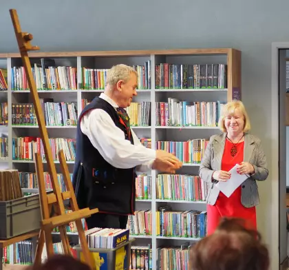 Pan Marek stoi koło sztalugi, a obok kobieta z kartką, za nimi regał z książkami.