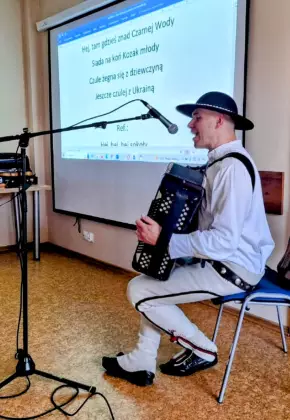 Góral Szczepan siedzi na krześle, gra na akordeonie i śpiewa do mikrofonu.