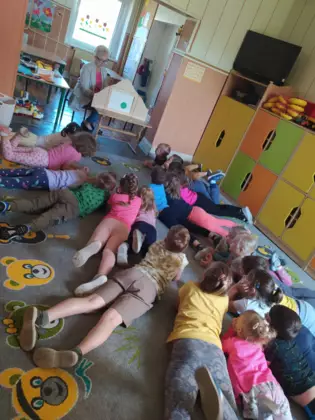 Dzieci leżą na dywanie a pani bibliotekarka czyta opowiadanie za pomocą teatrzyku kamishibai.