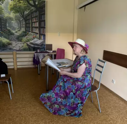 Maria Dziuk w kolorowej sukience siedzi przy stoliku i czyta wiersz.