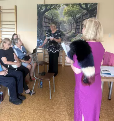 Grupa seniorów siedzi na sali. jedna z uczestniczek czyta wiersz.