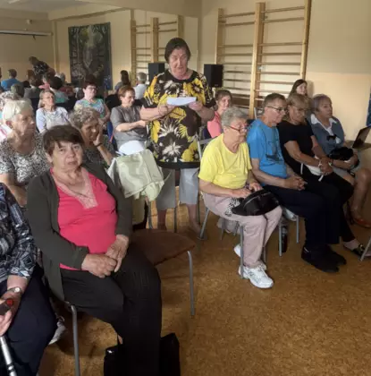 Grupa seniorów siedzi na sali. jedna z uczestniczek czyta wiersz.