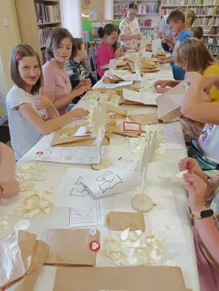 Dzieci siedzą przy stole i wyklejają statuetkę Orła Białego wiórkami osikowymi.