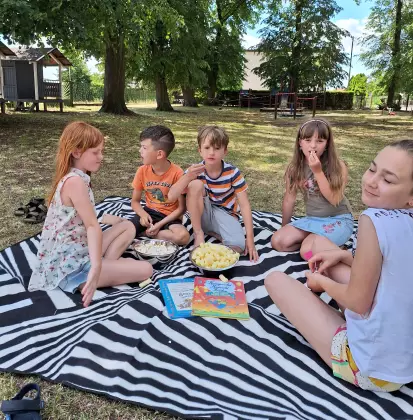Dzieci siedzą na kocu piknikowym, jedzą chrupki i słuchają czytanej bajki.