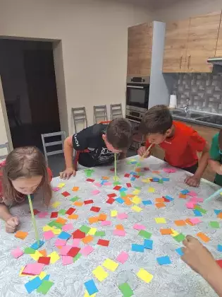 Dzieci bawią się w zbieranie kolorowych karteczek słomkami.