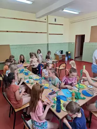 Dzieci siedzą przy stolikach i malują farbami talerzyki papierowe.