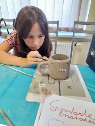 Dziewczyna siedzi przy stole i maluje farbą kubek ceramiczny.