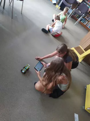 Dziewczyny jeżdżą samochodem złożonym z lego w tle dwóch chłopców ze swoimi pojazdami.