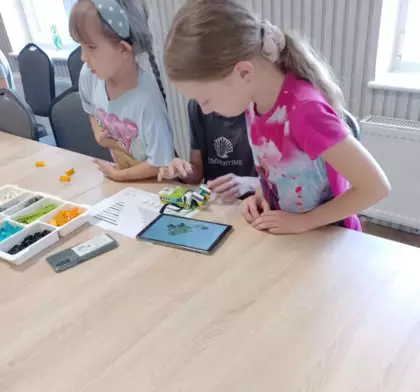 Dwie uczestniczki zajęć siedzą przy stolikach w bibliotece i budują pojazdy jeżdżące z klocków lego.