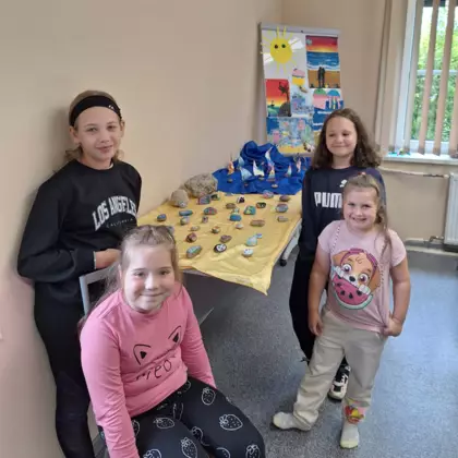 Wspólne zdjęcie po zajęciach. W tle mata morska na której pływają stateczki a obok znajdują się pomalowane przez dzieci na zajęciach kamienie.