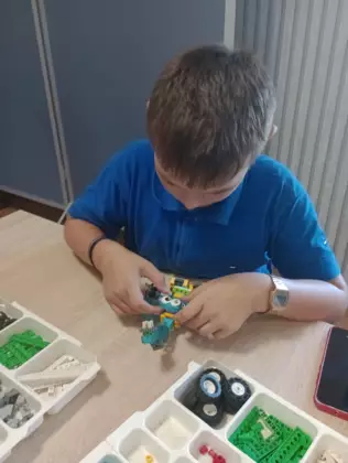 Dzieci siedzą przy stolikach w bibliotece i budują pojazdy z klocków lego..