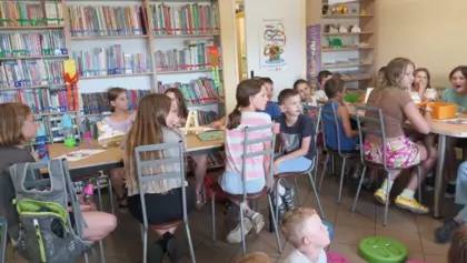 Dzieci siedzą przy stolikach i słuchają czytanej bajki przez bibliotekarkę.