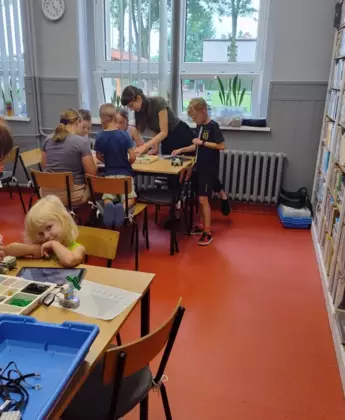Dzieci siedzą przy stolikach w bibliotece i budują pojazdy jeżdżące.