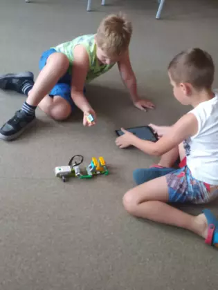 Dwóch chłopaków siedzi na podłodze i uruchamia pojazd złożony z klocków.