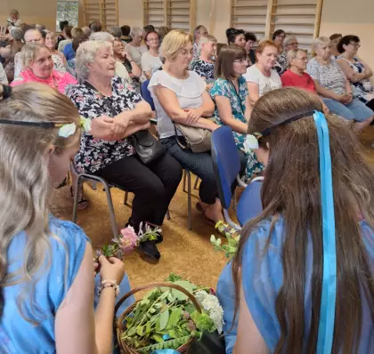 Dziewczynki wiją wianki a w tle siedzą uczestnicy spotkania.