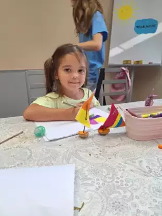 Dziewczynka maluje obrazek a przed nią stoją stateczki zrobione z łupinek po orzechach.