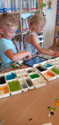 Uczestnicy zajęć budują przy stoliku w bibliotece pojazdy z klocków lego.