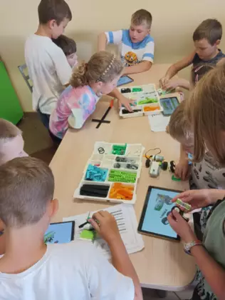 Uczestnicy zajęć budują przy stoliku w bibliotece pojazdy z klocków lego.