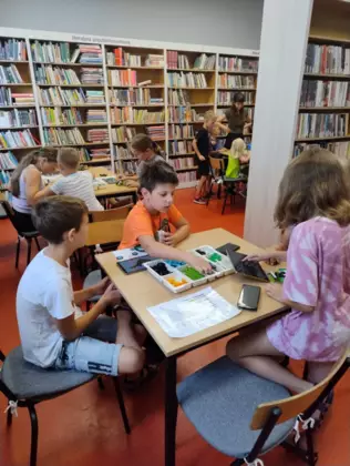 Dzieci siedzą przy stolikach w bibliotece i budują pojazdy jeżdżące.