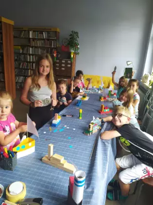 Uczestnicy zajęć pozują do zdjęcia siedząc przy stole w bibliotece ze swoimi pracami plastycznymi.