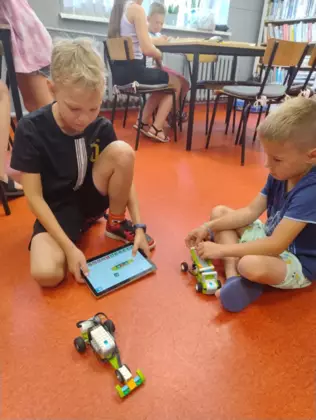 Uczestnicy jeżdżą zbudowanymi pojazdami po bibliotece.