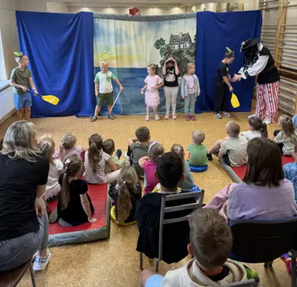 Dzieci oglądają teatrzyk – wśród widowni są przedszkolaki i starsze dzieci, które uważnie patrzą na występ i aktywnie w nim uczestniczą.