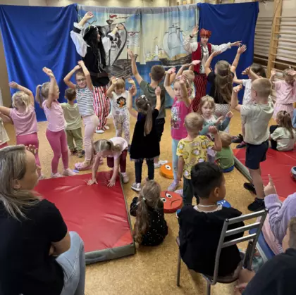 Dzieci oglądają teatrzyk – wśród widowni są przedszkolaki i starsze dzieci, które uważnie patrzą na występ i aktywnie w nim uczestniczą.
