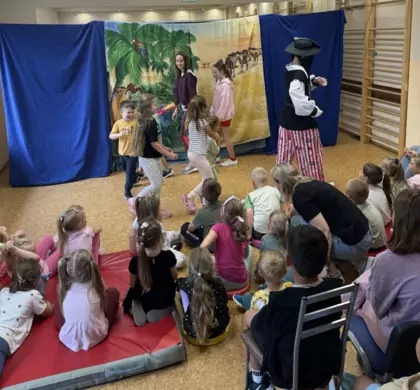 Dzieci oglądają teatrzyk – wśród widowni są przedszkolaki i starsze dzieci, które uważnie patrzą na występ i aktywnie w nim uczestniczą.