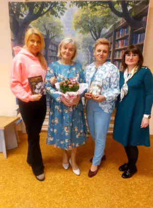 Cztery kobiety pozują do wspólnego zdjęcia podczas spotkania autorskiego w bibliotece. Jedna z nich trzyma bukiet kwiatów, a dwie prezentują książki autorki. W tle znajduje się dekoracja przedstawiająca regały z książkami i drzewa, nawiązująca do bibliotecznej atmosfery.