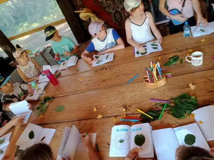 Na warsztatach dendrologicznych dzieci wykonują przy stole atlasy różnych gatunków drzew i krzewów. Zielone liście przyklejają do zeszytu.