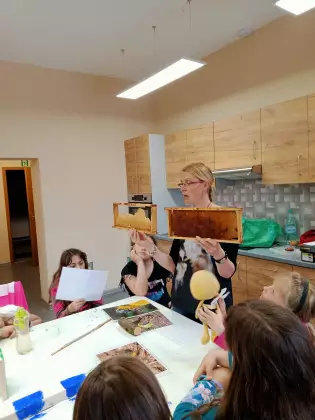 Na warsztatach przyrodniczych Pani przyrodnik stoi i trzyma dwa plastry miodu. Dzieci patrzą zaciekawione.