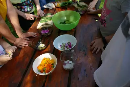 Na stole kilka miseczek z sałatkami z kolorowych kwiatów. Dookoła stołu stoją dzieci, jedno z dzieci kroi liście.