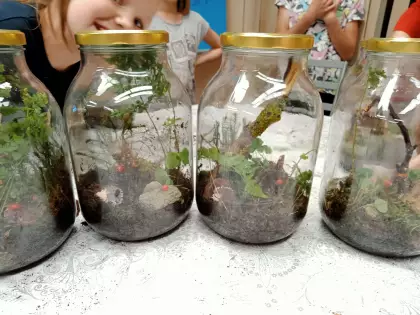 Na stole cztery duże zakręcone słoje z kompozycją leśną w środku. Dziewczynka patrzy z uśmiechem.