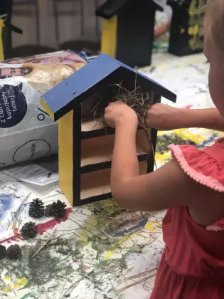 Dziewczynka przy stole wkłada siano do drewnianego domku dla owadów.