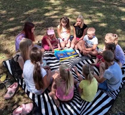Dzieci siedzą na kocu piknikowym i słuchają czytanej bajki.