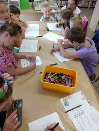 Dzieci siedzą przy stole i malują kredkami portret Aleksandra Fredry.