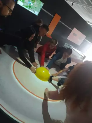 W ciemnym pomieszczeniu grupa ludzi dotyka model wnętrza Ziemi. Ręce wszystkich dotykają środka wnętrza Ziemi w formie żółtej półkuli.